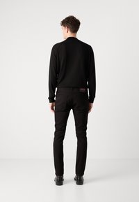 Haut noir à manches longues avec un col rond, associé à un pantalon noir slim et des chaussures noires, vu de dos. Détail logo rouge sur le pantalon.