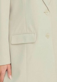 Ljusbeige kavaj med slagkrage, två knappar och en sidficka. Tyget har en mjuk textur med minimal detaljering.