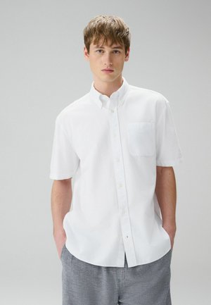 Junger Mann mit hellbraunen Haaren, der ein weißes kurzärmeliges Hemd mit Knöpfen und graue Hose trägt, steht mit den Händen in den Taschen.