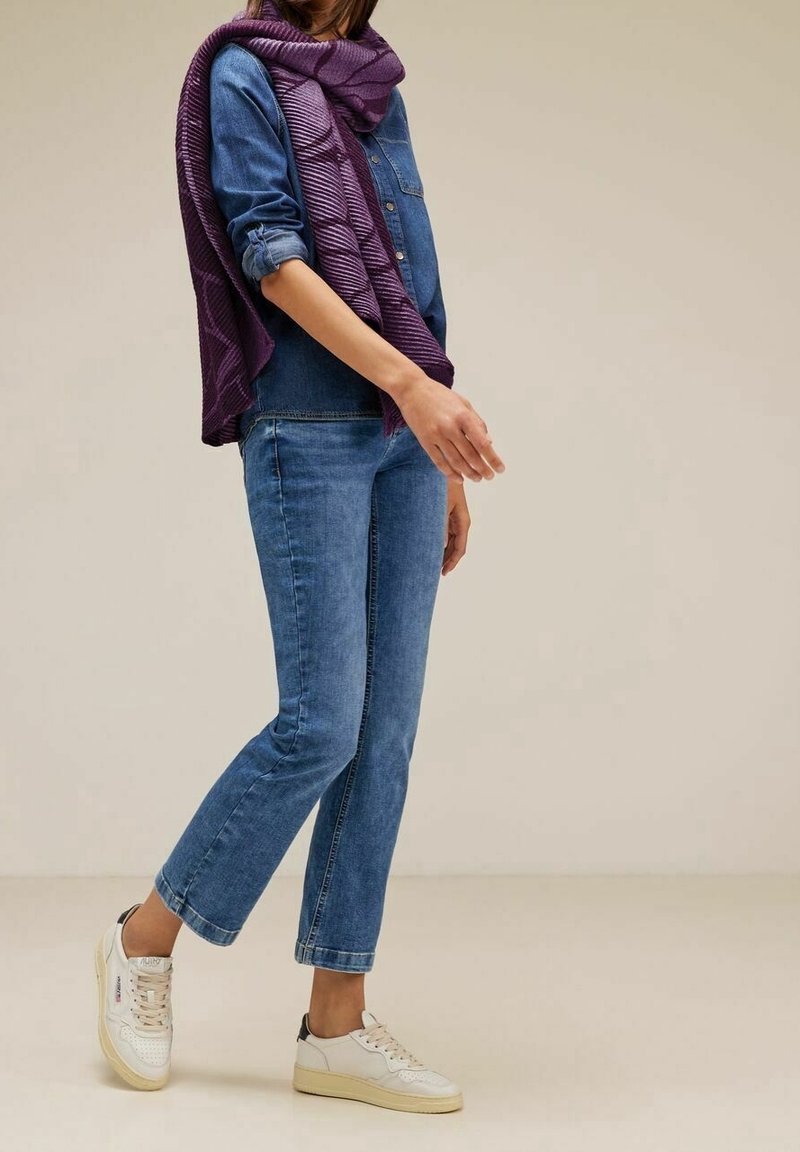 Femme portant une chemise en denim bleu et un jean avec une écharpe violette et des baskets blanches sur un fond uni.