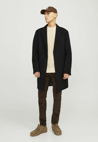 Abrigo de lana negro sobre un suéter texturizado beige, combinado con pantalones de pana marrones y zapatos de cordones color tostado. El modelo lleva una gorra marrón.