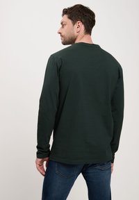 Ciemnozielona koszula z długim rękawem, wykonana z bawełny, z okrągłym dekoltem i prostym dołem. Model ma na sobie niebieskie dżinsy.