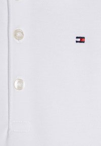 Valkoinen pikeepaita teksturoidusta kankaasta, jossa on kolmen napin nappilista, kirjailtu logo punaisena, valkoisena ja sinisenä sekä tasakudottu kaulus.