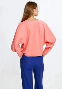 Koralowa bluza z luźnym krojem i szerokimi rękawami, w parze z niebieskimi spodniami. Gładka faktura i zaokrąglony dekolt widoczny od tyłu.