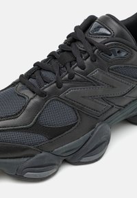 Scarpa sportiva nera realizzata con un mix di materiali in pelle e rete, forma curva, superficie testurizzata e un logo prominente sul lato.