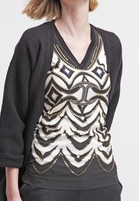 Černý kardigan přes bezrukávový vzorovaný top s geometrickými motivy v zlaté a modré na bílém pozadí. Měkká látka s výstřihem do V.