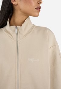 Béžová bundová mikina na zips s vysokým golierom, s textúrovanou tkaninou a rebrovým detailom. Biele logo "Pegador" vyšívané na prsiach.