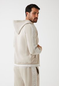 Lichtbeige ritshoodie met een capuchon, geribbelde manchetten en horizontale strepen. Gelaagde geprinte accenten op de mouwen. Zachte stof.