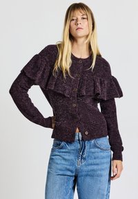 Dunkellila Strickjacke mit rüschendetail an der Vorderseite, Knopfverschluss und gerippten Bündchen, kombiniert mit hellblauen, hochtaillierten Jeans.