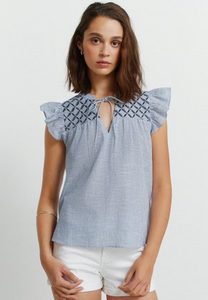 Ung kvinde iført en blå og hvidstribet ærmeløs top med broderet raglan og hvide shorts, stående mod en ensfarvet baggrund.