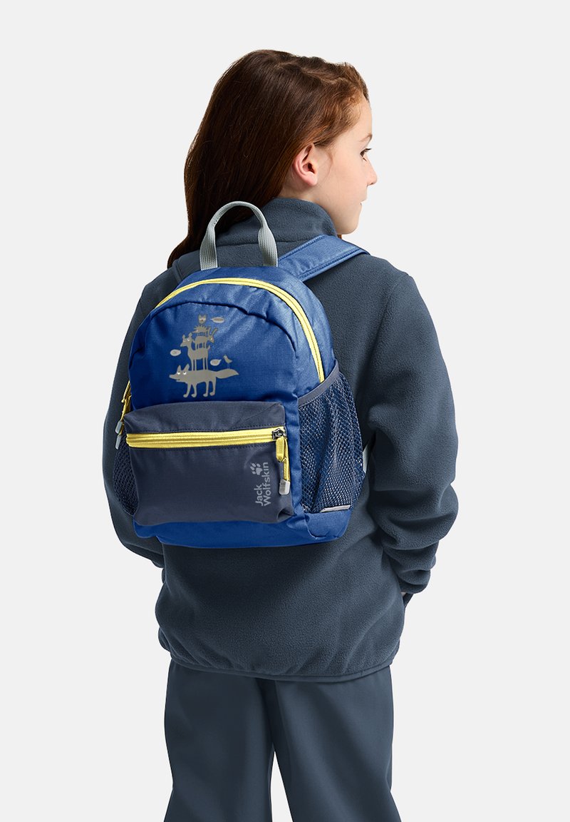 Παιδί πλάγια φορώντας μπλε φλις και μικρό μπλε σακίδιο πλάτης με κίτρινους φερμουάρ και σχέδιο με ζωάκια σε απλό φόντο.