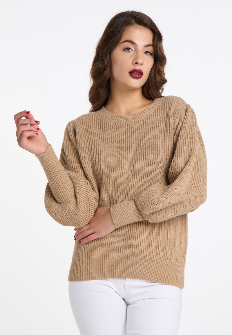 Beige kötött pulóver kerek nyakkal, bordázott mintával és enyhén puffasztott ujjal. Fehér nadrággal párosítva. Kezek összehúzva az előtérben.