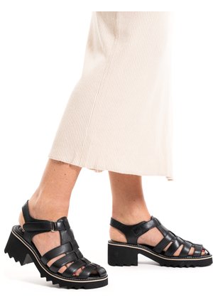 Pieds portant des sandales noires à talons épais avec plusieurs brides et une boucle, associées à un pantalon crème côtelé à jambes larges.