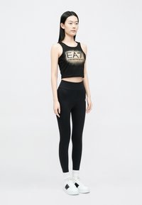 Fekete, kivágott top arany logóval, fekete, magasderekú leggingsszel párosítva. Fehér sportcipő fekete részletekkel. Egyszerű, testhez simuló kialakítás.
