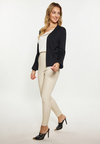 Cardigan nero a coste aperto davanti sopra una maglietta bianca, abbinato a pantaloni beige aderenti e mules neri a punta con accenti dorati.