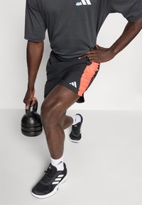 Un short de sport noir avec des panneaux latéraux orange, un t-shirt gris et des chaussures noires. Un kettlebell repose sur le sol à proximité.