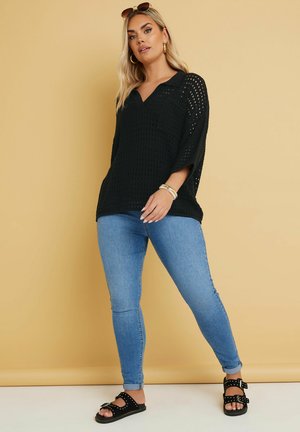 Schwarzes, offenes Strickoberteil mit V-Ausschnitt und kurzen, weiten Ärmeln, kombiniert mit hellblauen Skinny-Jeans und schwarzen Sandalen mit Nietenakzenten.