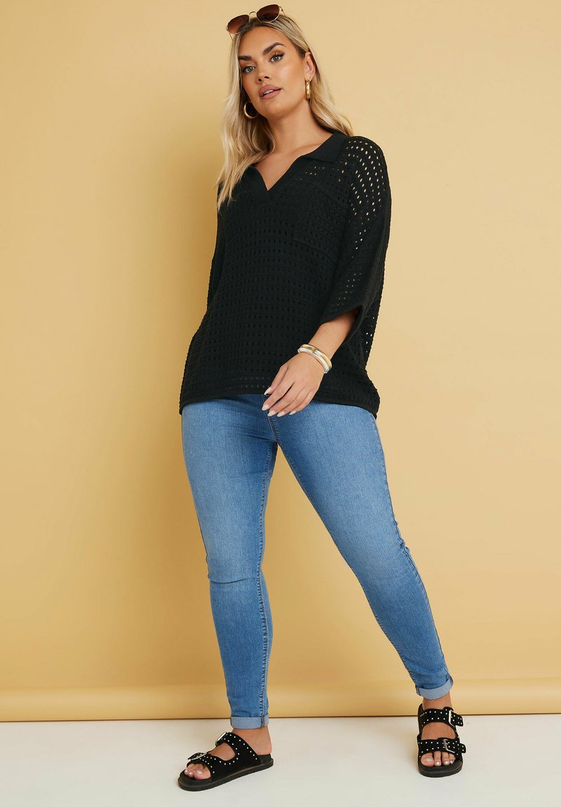 Schwarzes, offenes Strickoberteil mit V-Ausschnitt und kurzen, weiten Ärmeln, kombiniert mit hellblauen Skinny-Jeans und schwarzen Sandalen mit Nietenakzenten.