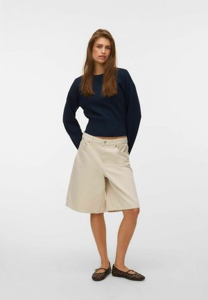 Jeune femme aux longs cheveux bruns portant un pull bleu marine foncé, un short large beige jusqu'aux genoux et des ballerines tissées marron foncé, debout les mains derrière le dos.