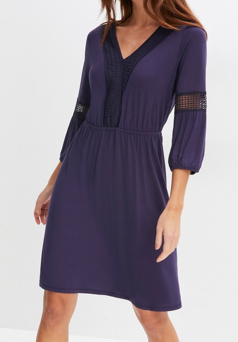 Femme portant une robe violette longueur genou avec manches 3/4, panneaux en dentelle et col en V avec bordure en dentelle assortie.