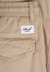 Bolsillo de pantalón beige con cintura elástica, trabilla para cinturón y etiqueta blanca cosida, con la inscripción "Reell est. 1997" en fuente de letra cursiva negra.