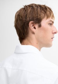 Weißes Hemd mit strukturiertem Kragen, glatter Textur und klaren Linien. Das kurze, zerzauste Haar des Models ist von der Profilansicht sichtbar.
