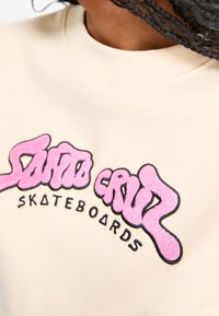 Felpa beige con un logo testurizzato rosa e nero che recita "Santa Cruz Skateboards". Presenta un collo rotondo e una consistenza di tessuto morbido.