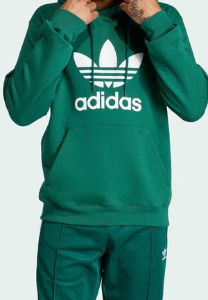 Person iført grøn Adidas hættetrøje og matchende grønne Adidas bukser, hænder justerer snorene på hættetrøjen, synlig tatovering på hånden.
