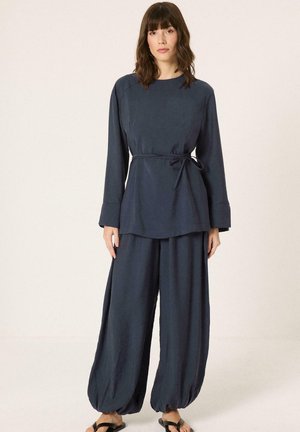 Γυναίκα στέκεται φορώντας φαρδιά μπλε μπλούζα με δέσιμο στο πλάι και ταιριαστό φαρδύ παντελόνι, συνδυασμένα με μαύρα σανδάλια σε απλό ανοιχτό φόντο.