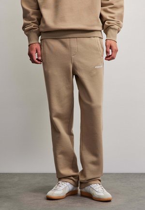 Man met beige joggingbroek en bijpassend sweatshirt, staand, met witte en beige sneakers op een grijze vloer tegen een effen muur.