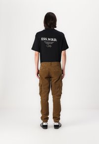 Schwarzes T-Shirt mit kurzen Ärmeln und weißen Text auf dem Rücken, kombiniert mit braunen Kordhosen im Cargo-Stil mit Taschen und schmal zulaufenden Säumen.