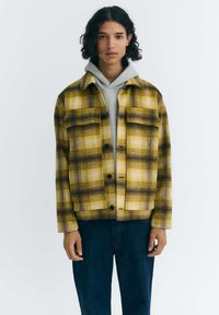 Gelb-braune kariert Jacke mit zwei Fronttaschen, Knopfverschluss und weichem Material, getragen über einem grauen Hoodie und dunklen Jeans.