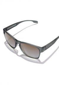 Ochelari de soare dreptunghiulari de culoare gri închis cu lentile nuanțate gradient și logo-ul "HAWKERS" pe braț, proiectând o umbră pe un fundal alb.