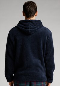 Námořnická modrá fleecová mikina s texturovaným, huňatým povrchem, s kapucí na šňůrku a volným střihem, viděná zezadu.