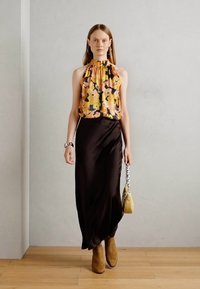 Kvinde, der går indendørs i en gul blomsteret halterneck top, lang sort nederdel, brune støvler, hoops øreringe og bærer en lille gul håndtaske.