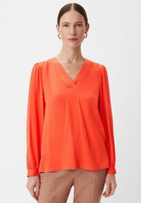 V-Ausschnitt, langärmelige Bluse aus leuchtend orangefarbenem Satinstoff. Mit gerafften Ärmeln und lockerer Passform. Kombiniert mit gemusterten Hosen.