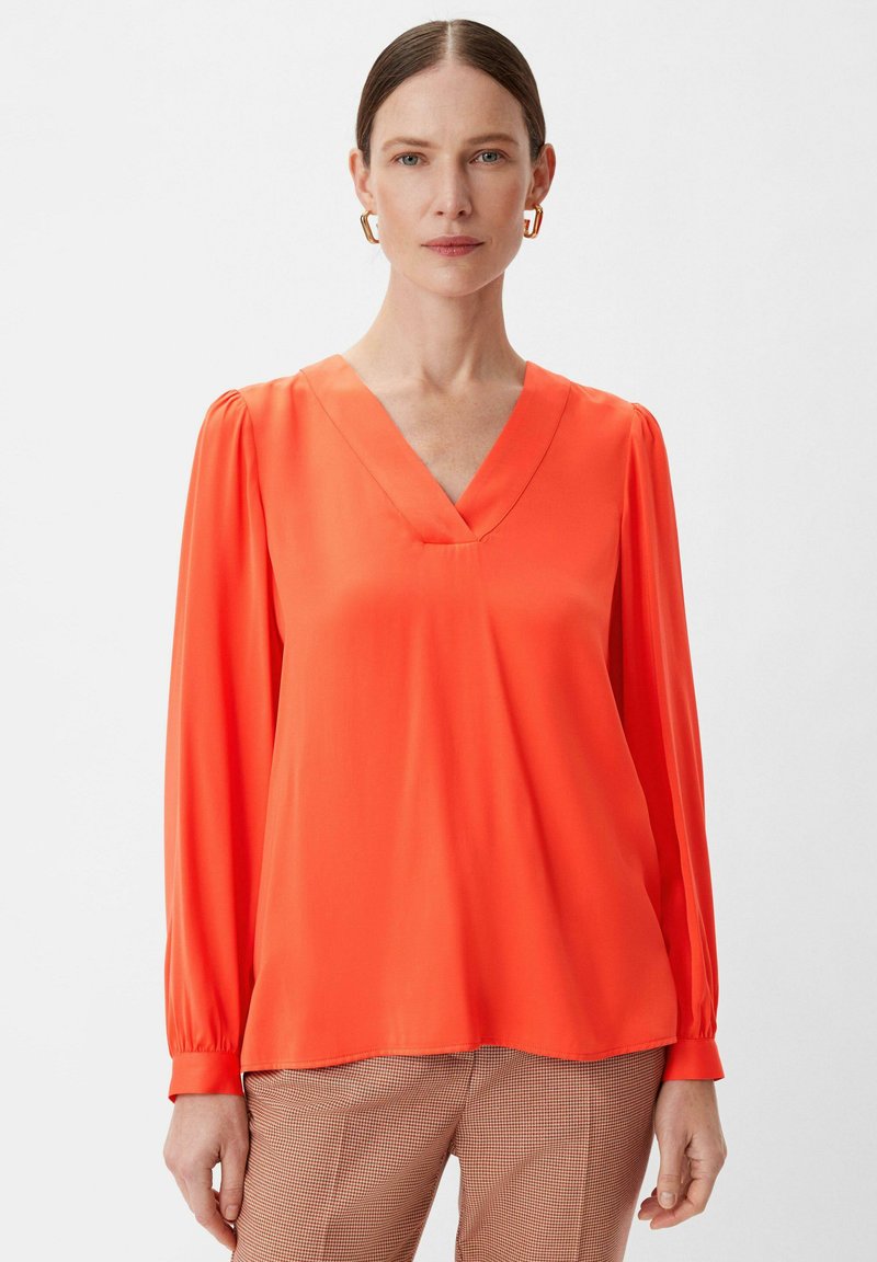 V-Ausschnitt, langärmelige Bluse aus leuchtend orangefarbenem Satinstoff. Mit gerafften Ärmeln und lockerer Passform. Kombiniert mit gemusterten Hosen.