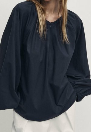 Femme portant un chemisier noir ample à manches longues avec poignets froncés et un pantalon clair sur un fond uni.