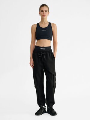 Młoda kobieta stojąca prosto, ubrana w czarny krótki top, czarne spodnie jogger typu cargo i czarne buty sportowe na jednolitym białym tle.