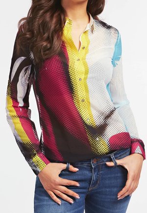 Femme portant une chemise à manches longues boutonnée avec un motif abstrait multicolore à pois et un jean bleu, mains reposant près des poches.