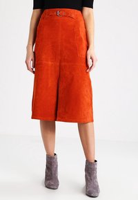 Nainen, jolla on polvipituinen oranssi nahkahelma, jossa on läpällisiä taskuja, yhteensopiva vyö, musta paita ja harmaat nahkaiset nilkkurit.