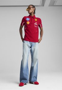 Tricou polo roșu cu logo-uri, accente albe și guler, asortat cu blugi evazați de culoare bleu deschis și pantofi roșii. Model care poartă ochelari de soare supradimensionați.