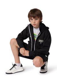 Sudadera negra con cremallera y detalles blancos, combinada con una camiseta gráfica y pantalones cortos negros. El calzado son zapatillas blancas con detalles negros.