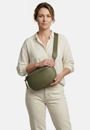 Frau in cremefarbenem Hemd und Hose, die eine olivgrüne Umhängetasche mit breitem Riemen und Reißverschluss hält, stehend vor einem weißen Hintergrund.