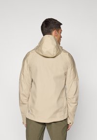 Veste légère beige avec capuche, présentant un design épuré et des coutures visibles. Agrémentée d'une petite étiquette de logo à l'ourlet.