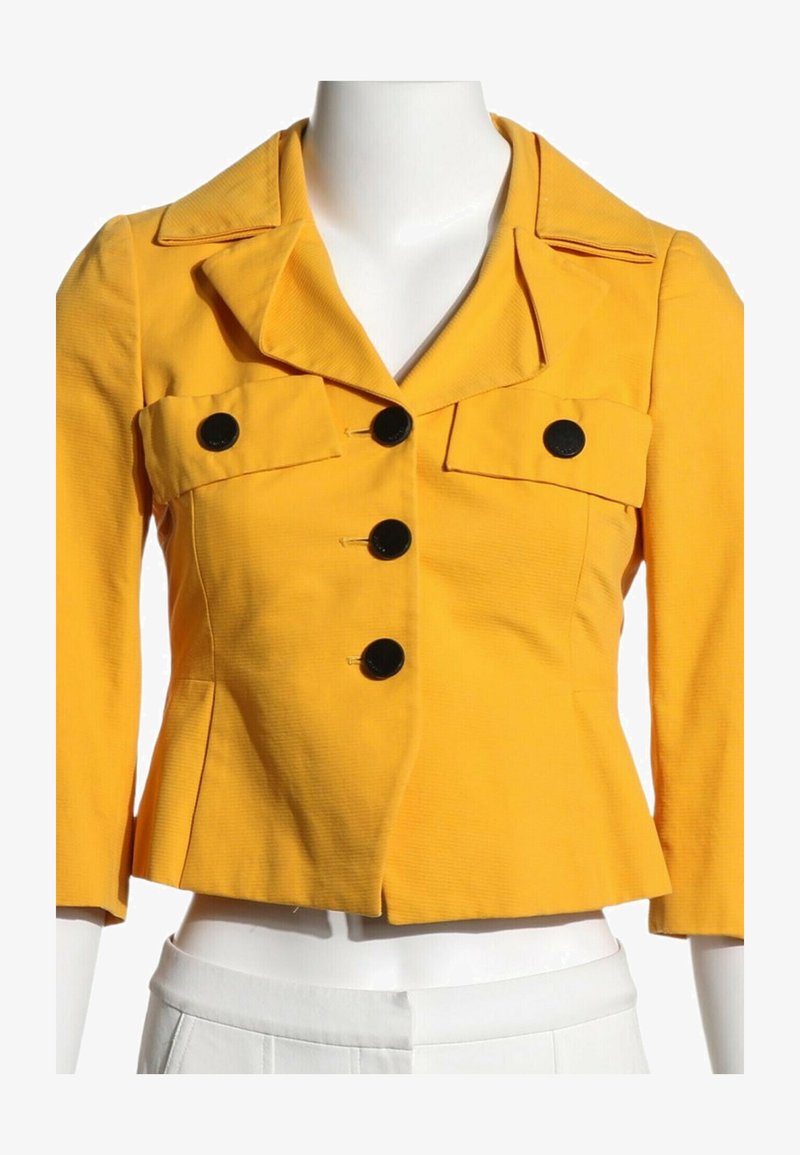 Veste courte jaune avec larges revers, trois grands boutons noirs sur le devant et deux poches poitrine boutonnées sur un mannequin blanc.
