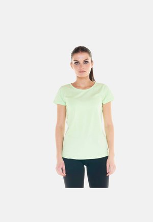 Donna con capelli scuri raccolti in una coda, in piedi, indossa una maglietta verde chiaro a maniche corte e pantaloni scuri, rivolta in avanti su uno sfondo uniforme.