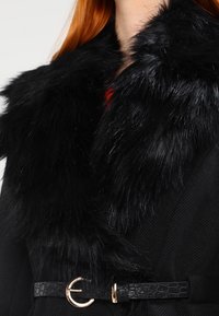 Svart kappa med en krage i konstpäls, som har en texturerad tyg och ett svart bälte med krokodilsmönster samt en metallisk cirkulär spänne.