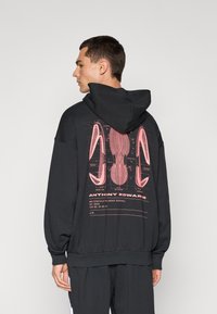 Sudadera con capucha negra que presenta un gran gráfico rosa en la parte posterior, mostrando detalles de diseño de zapatos. Tejido suave con un ajuste relajado.