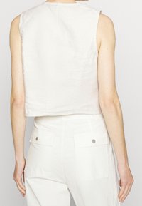 Kraftig ärmlös topp i ljusbeige linne, med en rak fåll och rena linjer, kombinerad med vita högmidjade byxor med fickor.